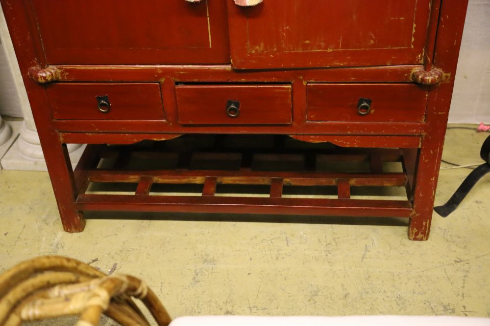 A Chinese two-door brass-mounted red-lacquered cupboard fitted three short drawers over an open shelf, width 110cm, depth 60cm, height